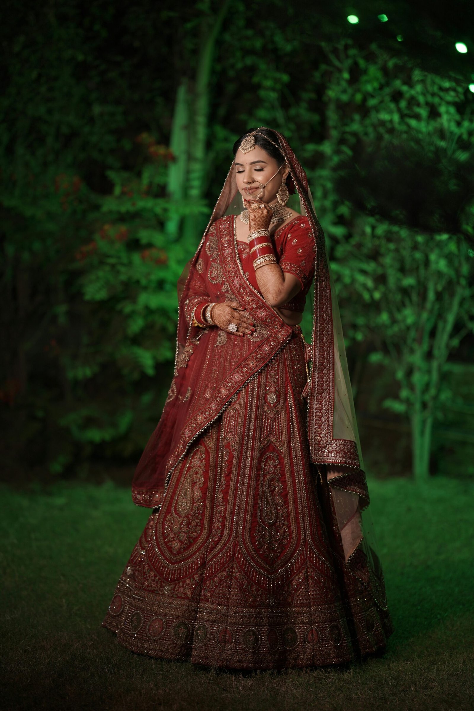 A beautiful South Asian bride poses gracefully in traditional red bridal attire outdoors.