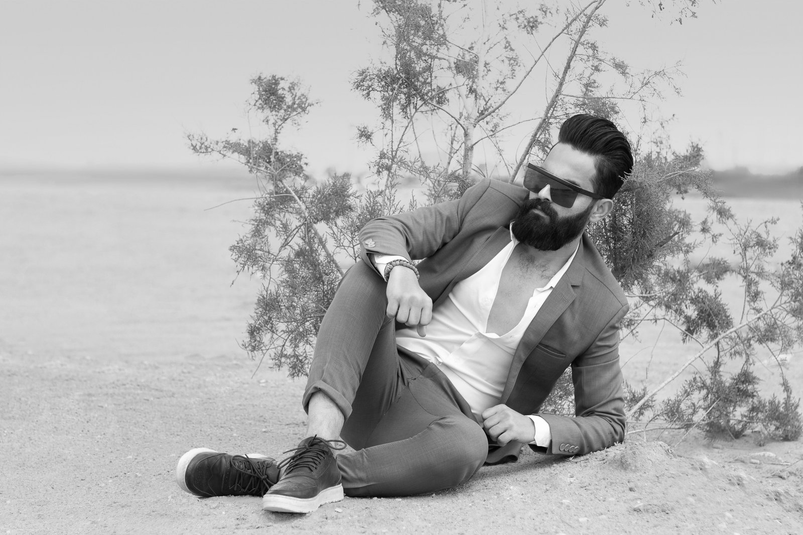Bearded man in a suit with sunglasses relaxes outdoors in grayscale style.