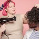 Female hairdresser with dyed hair drying hair of African American male model with Afro hairstyle against pink background